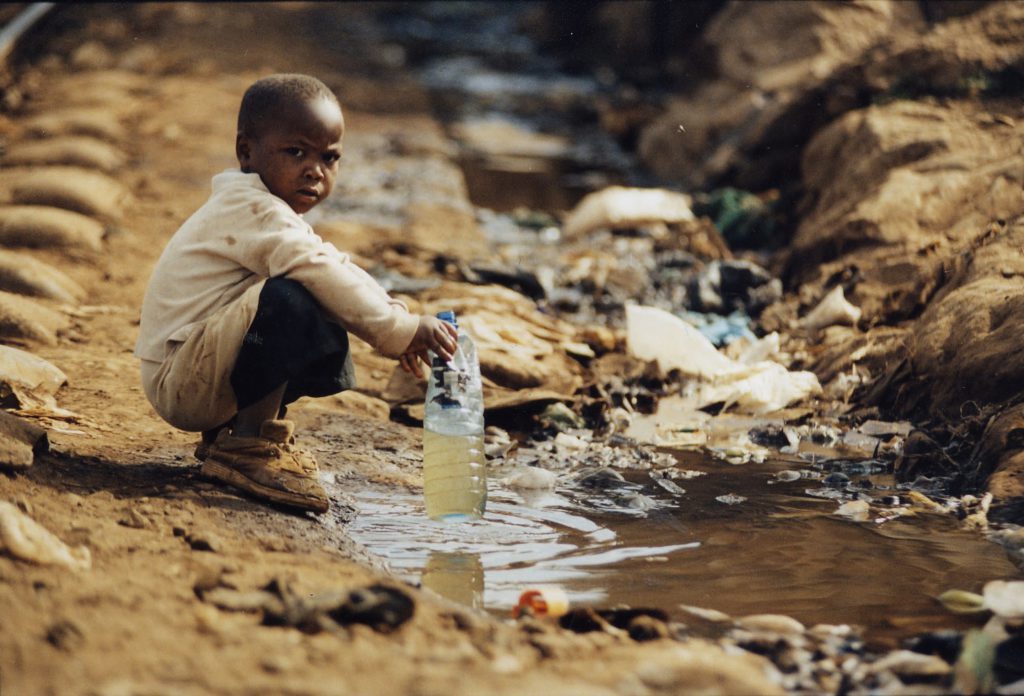 kid sitting with a water bottle in hand beside dirty water